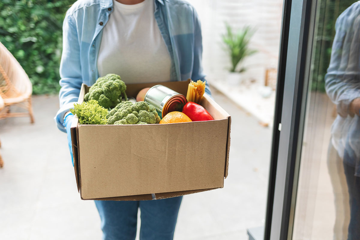 A woman smiles while holding a box filled with fresh vegetables and colorful fruits, ready for a healthy meal.
