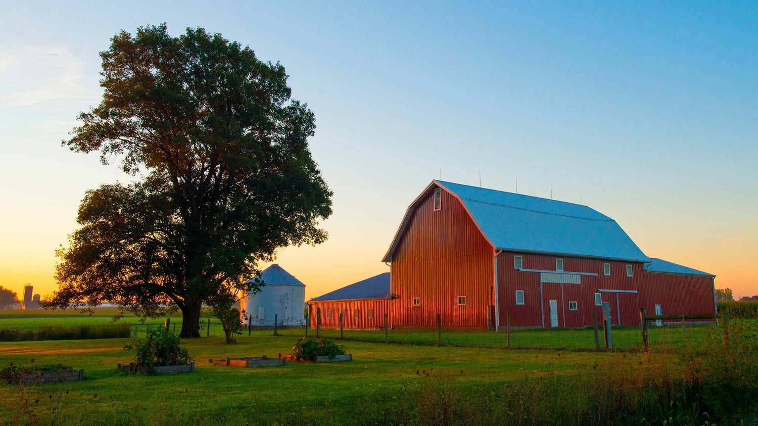 A vibrant red barn and silo silhouetted against a stunning sunset sky, casting warm hues of orange and pink.