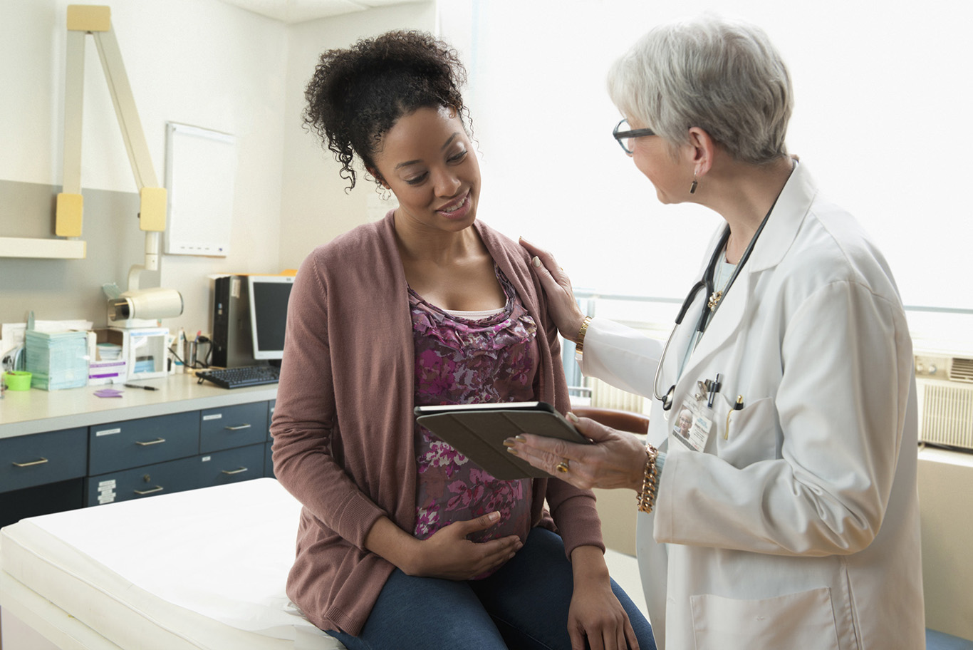 A pregnant woman cradles her belly as her doctor shows her information on a clipboard.
